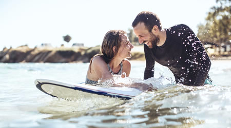 A couple in the water, laughing on a surfboard