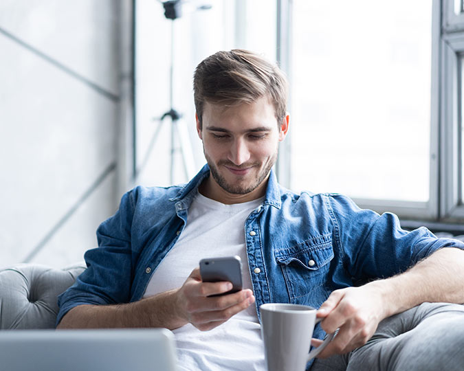 Man sitting on couch, looking at his phone while drinking coffee