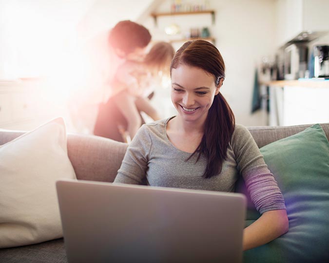 Woman working on laptop at home while son and child play behind her